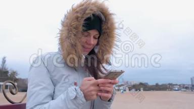 穿着白色羽绒服的女人坐在海滩上的长凳上，在手机上输入一条信息。